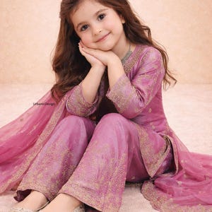 May include: A young girl wearing a pink embroidered outfit, including a top, pants, and a matching scarf. The outfit has gold embroidery. She is wearing silver flats. The image has the text "UrbanicDesign".
