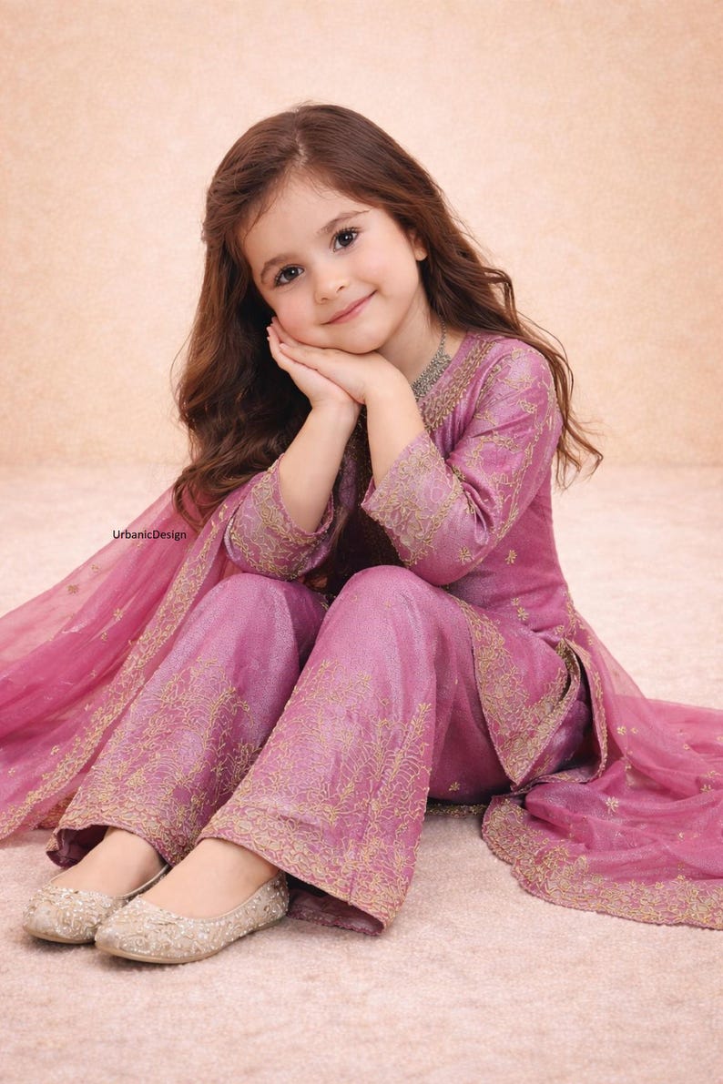 May include: A young girl wearing a pink embroidered outfit, including a top, pants, and a matching scarf. The outfit has gold embroidery. She is wearing silver flats. The image has the text "UrbanicDesign".