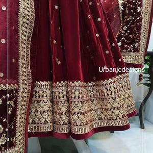 May include: A burgundy velvet garment with intricate gold embroidery. The hemline and edges feature detailed patterns. The garment is displayed on a mannequin, showcasing the rich color and texture. The text "Urbanicdesigns" is visible.