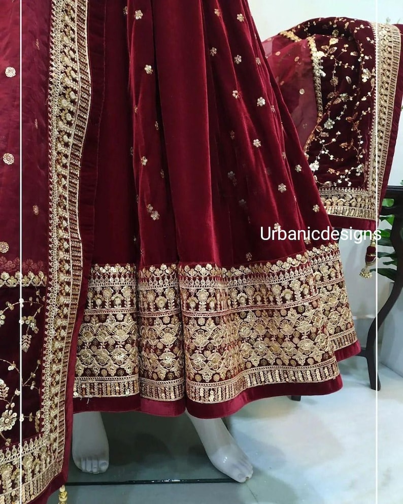 May include: A burgundy velvet garment with intricate gold embroidery. The hemline and edges feature detailed patterns. The garment is displayed on a mannequin, showcasing the rich color and texture. The text "Urbanicdesigns" is visible.