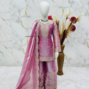 May include: A pink, embroidered salwar kameez set on a mannequin. The set includes a long-sleeved tunic, wide-legged pants, and a sheer scarf. The outfit is embellished with gold embroidery. A vase of flowers is in the background.