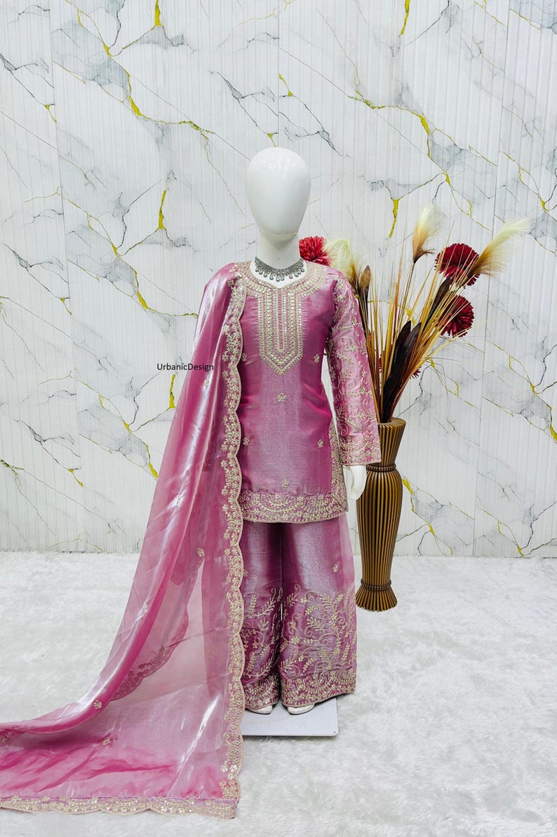 May include: A pink, embroidered salwar kameez set on a mannequin. The set includes a long-sleeved tunic, wide-legged pants, and a sheer scarf. The outfit is embellished with gold embroidery. A vase of flowers is in the background.