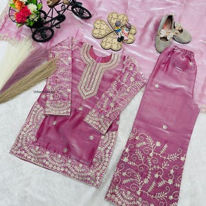 May include: A magenta-colored, embroidered salwar kameez set. The long-sleeved tunic and matching pants feature intricate gold threadwork. The set is displayed on a pink fabric, with decorative items like a miniature bicycle and shoes.