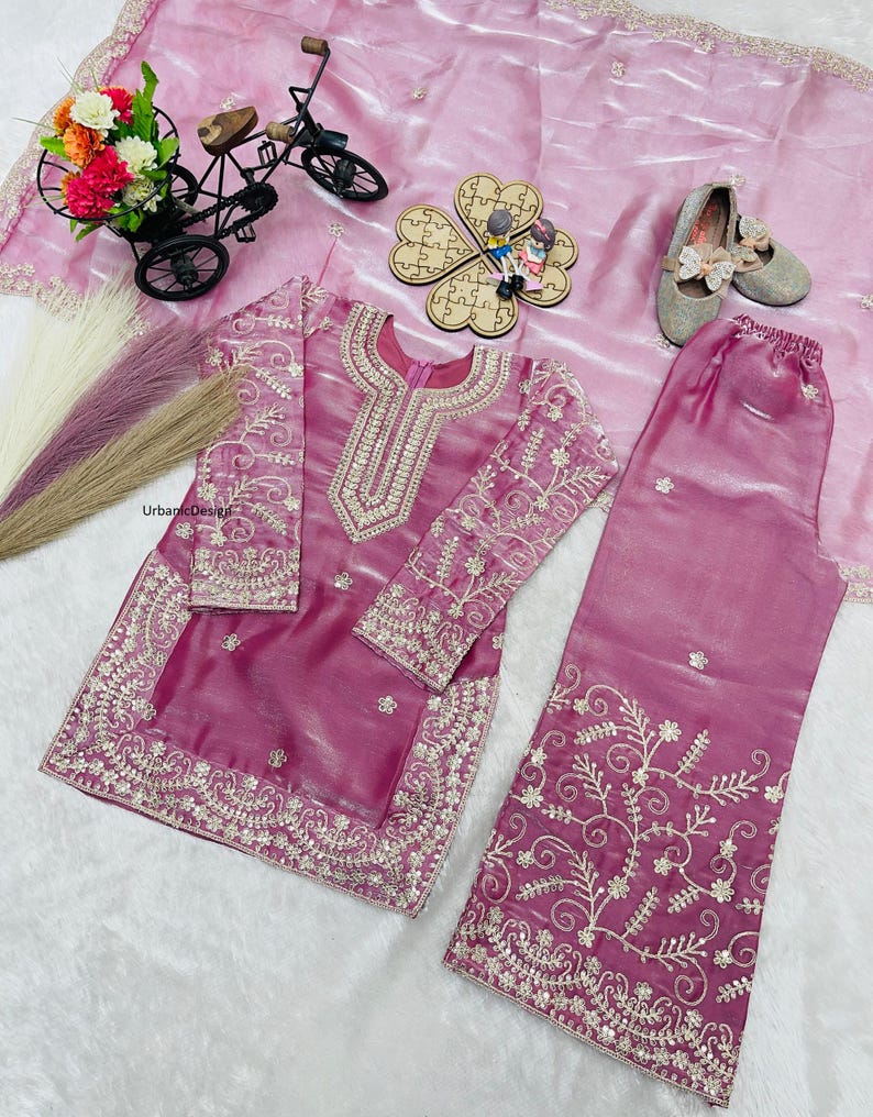 May include: A magenta-colored, embroidered salwar kameez set. The long-sleeved tunic and matching pants feature intricate gold threadwork. The set is displayed on a pink fabric, with decorative items like a miniature bicycle and shoes.