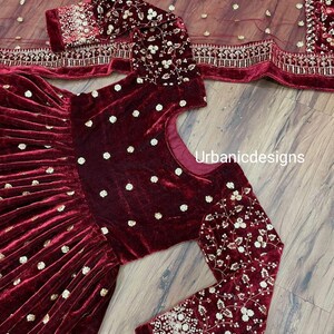 May include: A burgundy velvet dress with long sleeves and a matching sheer scarf. The dress features gold embroidered floral patterns on the sleeves and bodice, with small gold dots scattered across the fabric. The text "Urbanicdesigns" is visible.