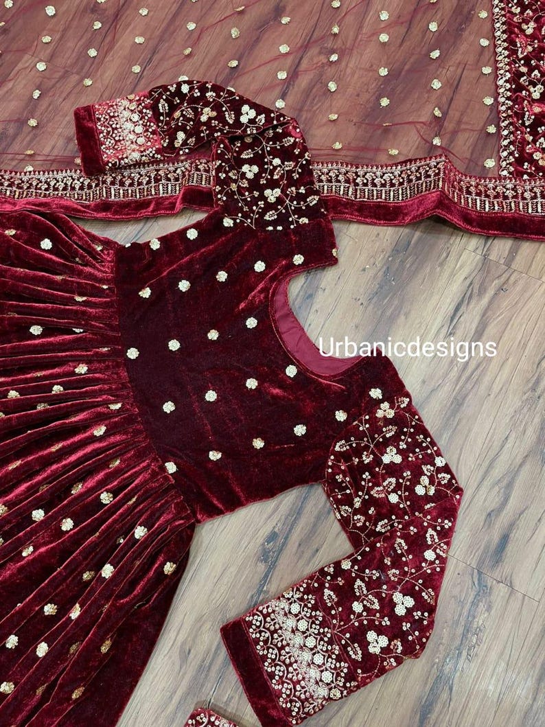 May include: A burgundy velvet dress with long sleeves and a matching sheer scarf. The dress features gold embroidered floral patterns on the sleeves and bodice, with small gold dots scattered across the fabric. The text "Urbanicdesigns" is visible.