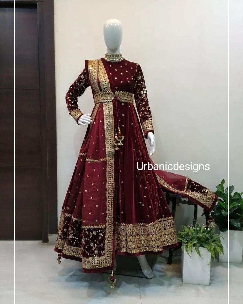 May include: A burgundy velvet gown with gold embroidery and embellishments. The long-sleeved dress features a high neckline, a fitted bodice, and a flowing skirt. The text "Urbanicdesigns" is visible on the dress.