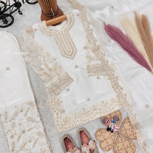 May include: A white embroidered outfit with matching pants and a sheer scarf. The outfit features gold embellishments and is paired with matching shoes with bow accents. A decorative wooden puzzle and a miniature bicycle are also present.