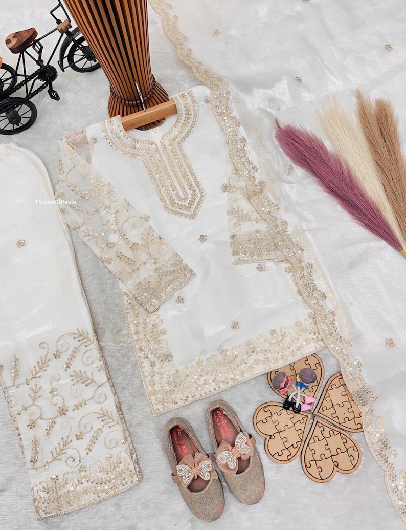 May include: A white embroidered outfit with matching pants and a sheer scarf. The outfit features gold embellishments and is paired with matching shoes with bow accents. A decorative wooden puzzle and a miniature bicycle are also present.