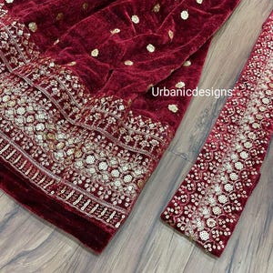 May include: A close-up of a burgundy velvet fabric with gold embellishments. The fabric has a floral embroidered border and scattered gold sequins. A matching sleeve is visible on the right side. The text "Urbanicdesigns" is visible on the fabric.