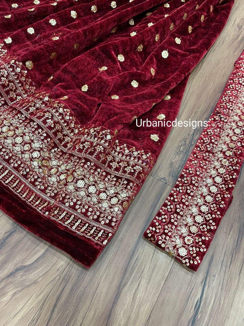May include: A close-up of a burgundy velvet fabric with gold embellishments. The fabric has a floral embroidered border and scattered gold sequins. A matching sleeve is visible on the right side. The text "Urbanicdesigns" is visible on the fabric.