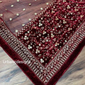 May include: A burgundy velvet fabric with intricate gold floral embroidery and sequin detailing. A sheer burgundy overlay is scattered with gold flower-shaped embellishments. The edge is trimmed with a thick, velvet border. The text "Urbanicdesigns" is visible.