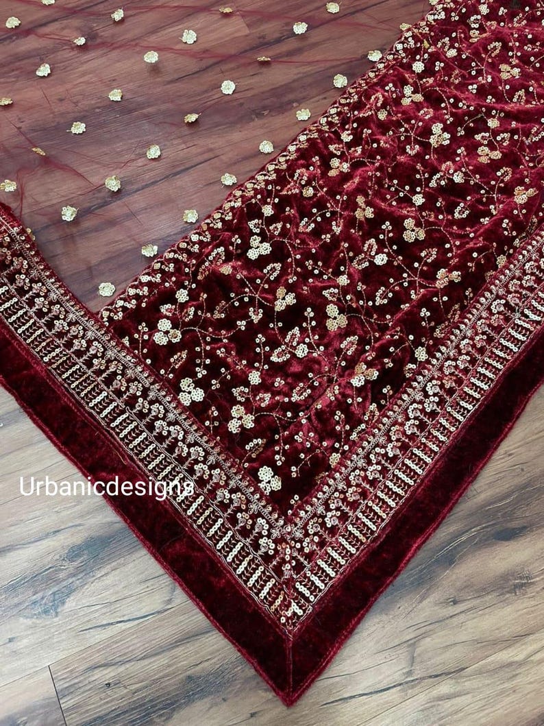 May include: A burgundy velvet fabric with intricate gold floral embroidery and sequin detailing. A sheer burgundy overlay is scattered with gold flower-shaped embellishments. The edge is trimmed with a thick, velvet border. The text "Urbanicdesigns" is visible.