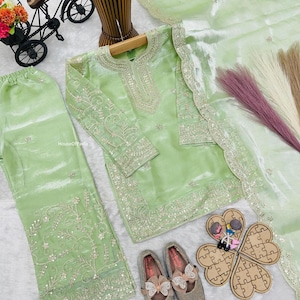 May include: A light green, embroidered salwar kameez set with matching pants and a sheer dupatta. The set includes a pair of gray shoes with bow accents. A decorative bicycle and a wooden puzzle are also present.