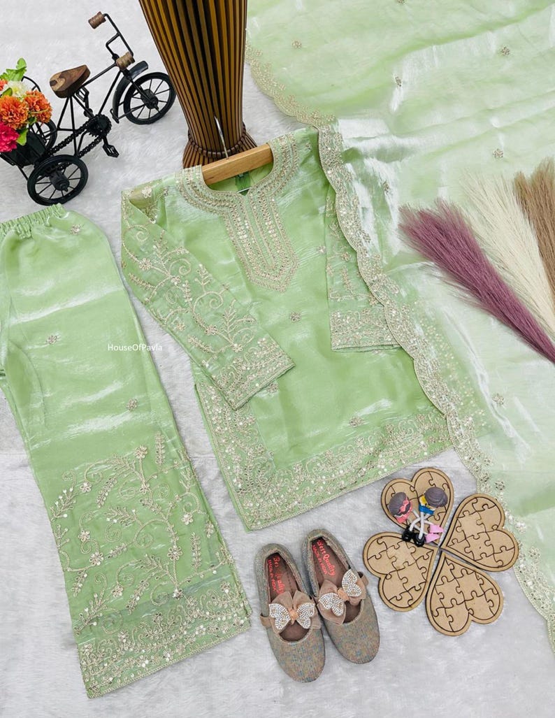 May include: A light green, embroidered salwar kameez set with matching pants and a sheer dupatta. The set includes a pair of gray shoes with bow accents. A decorative bicycle and a wooden puzzle are also present.