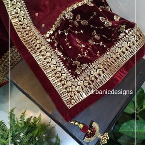 May include: A burgundy velvet shawl with intricate gold embroidery and sequin detailing. The shawl features a gold border and a decorative tassel. The design includes floral and geometric patterns, creating a luxurious aesthetic.