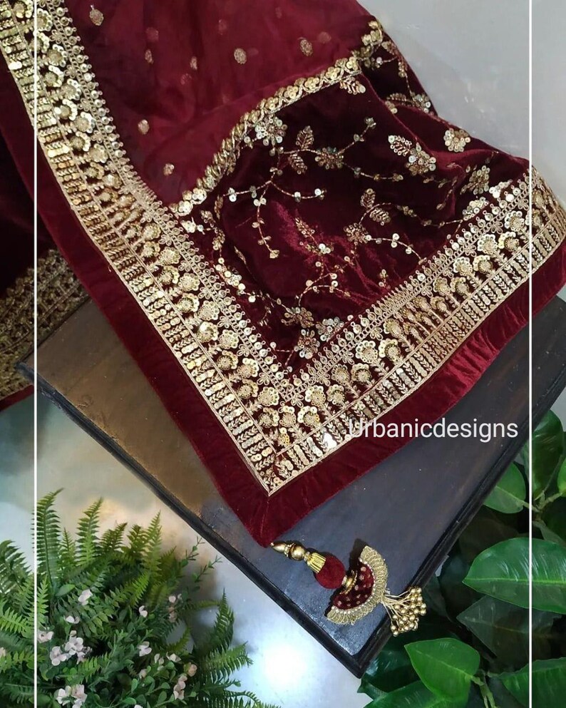 May include: A burgundy velvet shawl with intricate gold embroidery and sequin detailing. The shawl features a gold border and a decorative tassel. The design includes floral and geometric patterns, creating a luxurious aesthetic.