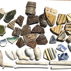 Puede incluir: Una colección de fragmentos y trozos de cerámica antigua, incluyendo piezas de pipas de cerámica, en varios colores como verde, marrón, azul y amarillo. Los fragmentos muestran patrones y texturas, sugiriendo un tema histórico.
