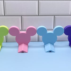 May include: Four Mickey Mouse head shaped wall hooks in different colors: green, pink, light blue, and purple. The hooks are mounted on a white tiled wall.