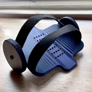 May include: A blue and black exercise wheel with a textured surface and black straps. The wheel has two black circular ends and a blue center with a series of small holes. The straps are made of a black, woven material.