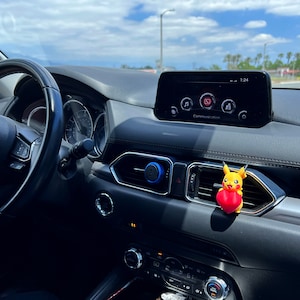 May include: The interior of a car with a black steering wheel, a black dashboard, and a touchscreen display showing the time 1:34. A yellow Pikachu character with a red heart is hanging from the air vent.