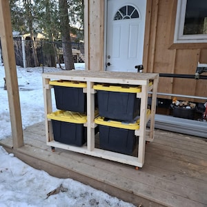 May include: A wooden storage unit with four black bins with yellow lids. The unit is on wheels and sits on a wooden deck. The background includes a white door and a snowy outdoor scene.