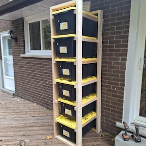 May include: A wooden storage rack with six black plastic bins with yellow lids. The rack is constructed from light-colored wood and is placed outdoors on a wooden deck. Two power drills are visible on the deck.