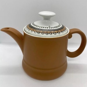 以下が含まれることがあります： 縁に白黒の幾何学模様が施された、茶色の陶器製のティーポット。蓋は白く、取っ手と注ぎ口が付いている。