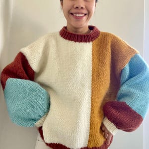 Può includere: Un maglione a blocchi di colore con girocollo e maniche lunghe. Il maglione presenta un pannello centrale color crema, con un pannello giallo senape e blu sul lato destro. Le maniche sono blu e color ruggine.
