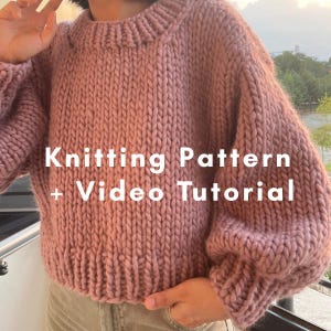 Op de afbeelding: Een grofgebreide trui in oudroze kleur met een ronde hals, lange mouwen met geribbelde boorden en een geribbelde zoom. De tekst Knitting Pattern + Video Tutorial staat in het wit over het voorpand van het kledingstuk heen.