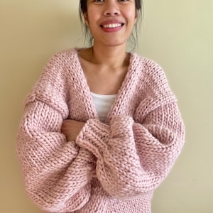 Könnte beinhalten: Eine grob gestrickte Strickjacke in zartem Pastellrosa. Das Kleidungsstück verfügt über einen tiefen V-Ausschnitt, lange Ärmel mit überschnittenen Schultern und ein durchgehendes, dickes und strukturiertes Strickmuster. Über einem weißen Oberteil.