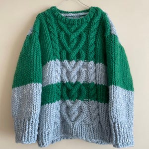 May include: A chunky knit sweater featuring horizontal stripes of vibrant green and light gray yarn. The garment has a crew neckline, long sleeves, and a prominent vertical cable knit pattern running down the center front of the torso.