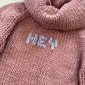 May include: A chunky knit sweater in a dusty rose color with a large, ribbed hood. The word "ME" is embroidered in light blue yarn on the front. The sweater has a thick, textured appearance, suggesting warmth and comfort.