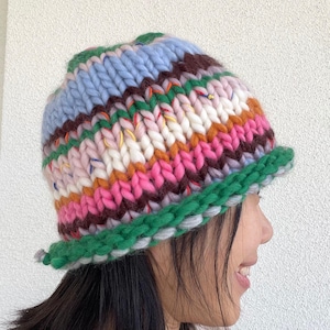 May include: A colorful, chunky knit beanie in shades of blue, pink, green, brown, and white. The hat features a rolled green brim and a textured, striped pattern. This handmade hat is a cozy winter accessory.