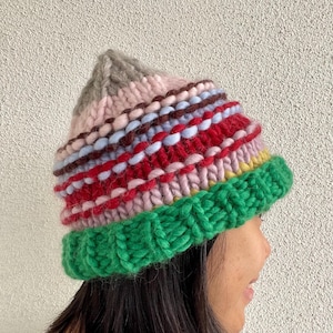 May include: A chunky knit beanie featuring horizontal stripes in various colors including green, yellow, pink, red, light blue, brown, and grey. The hat has a thick, ribbed green brim and a pointed crown, constructed from thick yarn.