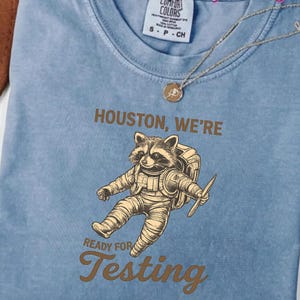 Peut inclure: T-shirt bleu clair Comfort Colors avec un dessin de raton laveur astronaute et le texte "Houston, We're Ready for Testing". Le t-shirt a une petite étiquette de taille et un collier doré.