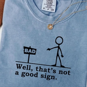以下が含まれることがあります： 「Well, that's not a good sign.」の文字と「BAD」と書かれた看板を指す棒人間のイラストがプリントされた、水色のComfort Colors Tシャツ。金のペンダントが付いたネックレスがかけられています。