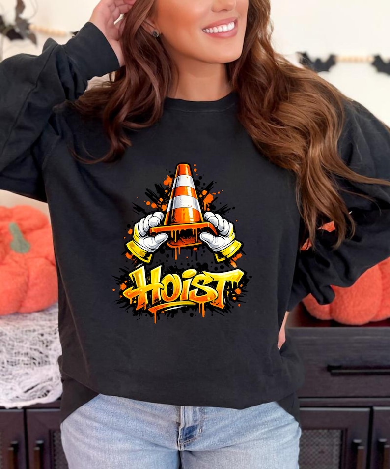 May include: Black long-sleeve shirt featuring a graphic of a traffic cone held by cartoon hands with yellow gloves. The word "Hoist" is written in orange graffiti-style lettering below the graphic. The shirt is a Halloween-themed design.