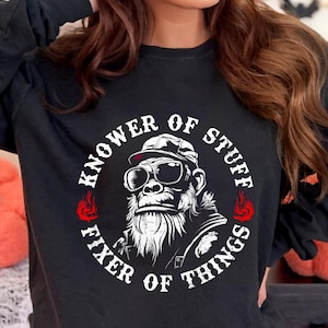 May include: Black long-sleeve shirt featuring a gorilla wearing sunglasses and a cap. The graphic includes the text "Knower of Stuff, Fixer of Things" in a circular design with red flame accents.