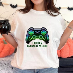 Peut inclure: T-shirt blanc avec un motif de manette de jeu vidéo colorée, des trèfles et le texte "LUCKY GAMER MODE" en vert. La manette présente un dégradé de bleu, vert et noir.