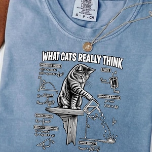 Peut inclure: Un t-shirt bleu clair avec un motif représentant un chat poussant un verre au bord d'une étagère, entouré d'équations de physique et du texte "WHAT CATS REALLY THINK". Le t-shirt possède un col rond et une étiquette Comfort Colors.
