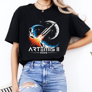 May include: Black t-shirt featuring a graphic of a rocket launching towards the moon, with the text "ARTEMIS II 2026" in white. The design includes blue, orange, and white elements. The shirt is a casual wear item.