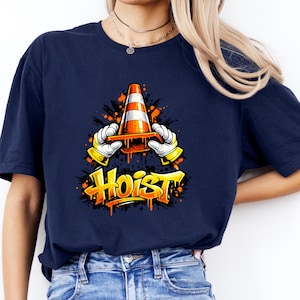 May include: Navy blue t-shirt featuring a graphic of a traffic cone held by cartoon hands with the word "Hoist" in yellow graffiti style. The design includes orange paint splatter effects.