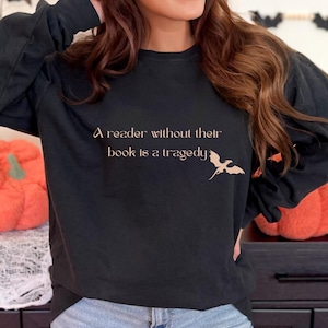 May include: Black long-sleeve sweatshirt with the quote "A reader without their book is a tragedy" in a copper color. A stylized dragon graphic is next to the text. The sweatshirt is worn by a person.