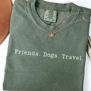 Peut inclure: Un t-shirt vert olive avec les mots "Friends. Dogs. Travel." imprimés en blanc. Un collier doré avec un pendentif circulaire repose sur le t-shirt. Le t-shirt est plié proprement.