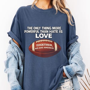 以下が含まれることがあります： 白文字で「THE ONLY THING MORE POWERFUL THAN HATE IS LOVE」と書かれたネイビーブルーのTシャツ。その下に「TOGETHER, WE ARE AMERICA」というテキストが入ったフットボールのグラフィックがあります。