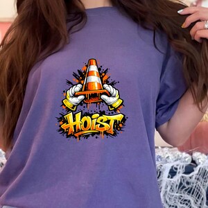 May include: A purple t-shirt featuring a graphic of a traffic cone held by gloved hands, with the word "Hoist" in graffiti-style lettering. The design includes orange and black accents.