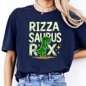 Puede incluir: Camiseta azul marino con el texto "RIZZA SAURUS REX" en fuente verde claro. Un dinosaurio verde con gafas de sol está en el centro. El diseño incluye acentos coloridos y estrellas.