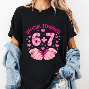 May include: Black t-shirt with the text "OFFICIAL TEENAGER 6+7" in pink, surrounded by pink hearts. The design includes a graphic of two hands with pink nail polish. The shirt is a casual style.