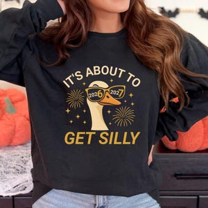 Peut inclure: Sweat-shirt noir avec un motif d'oie portant des lunettes avec les chiffres 2026 et 2027. Le texte "IT'S ABOUT TO GET SILLY" est imprimé en or. Le motif comprend également des feux d'artifice et des étoiles.